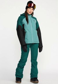 Groenblauw en zwart geïsoleerd sneeuwjack gecombineerd met groenblauwe broek. Voorzien van een ritssluiting aan de voorkant, capuchon en zwarte handschoenen. Inclusief een zwarte muts en skibril.