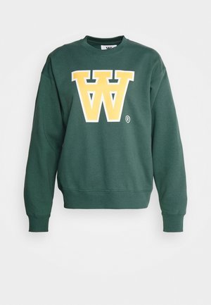 Henkilö, joka pitää päällään vihreää collegepaitaa, jossa on keltainen "W"-logo, ruudulliset housut ja valkoiset tennarit, seisoo ulkona metalliritilällä lähellä puita ja aitaa.