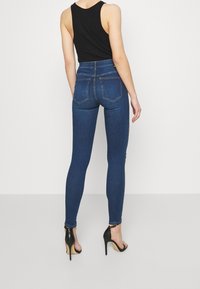 Jeans ajustados en denim azul oscuro, diseño de cintura alta, con bolsillos traseros, combinados con sandalias negras de tacón alto. Textura suave.