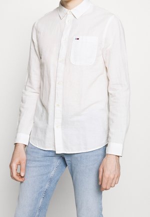 Biała koszula na długi rękaw z guzikami, kieszenią na piersi i małym logo, noszona z jasnoniebieskimi dżinsami, widok z przodu na tułów i ramiona.
