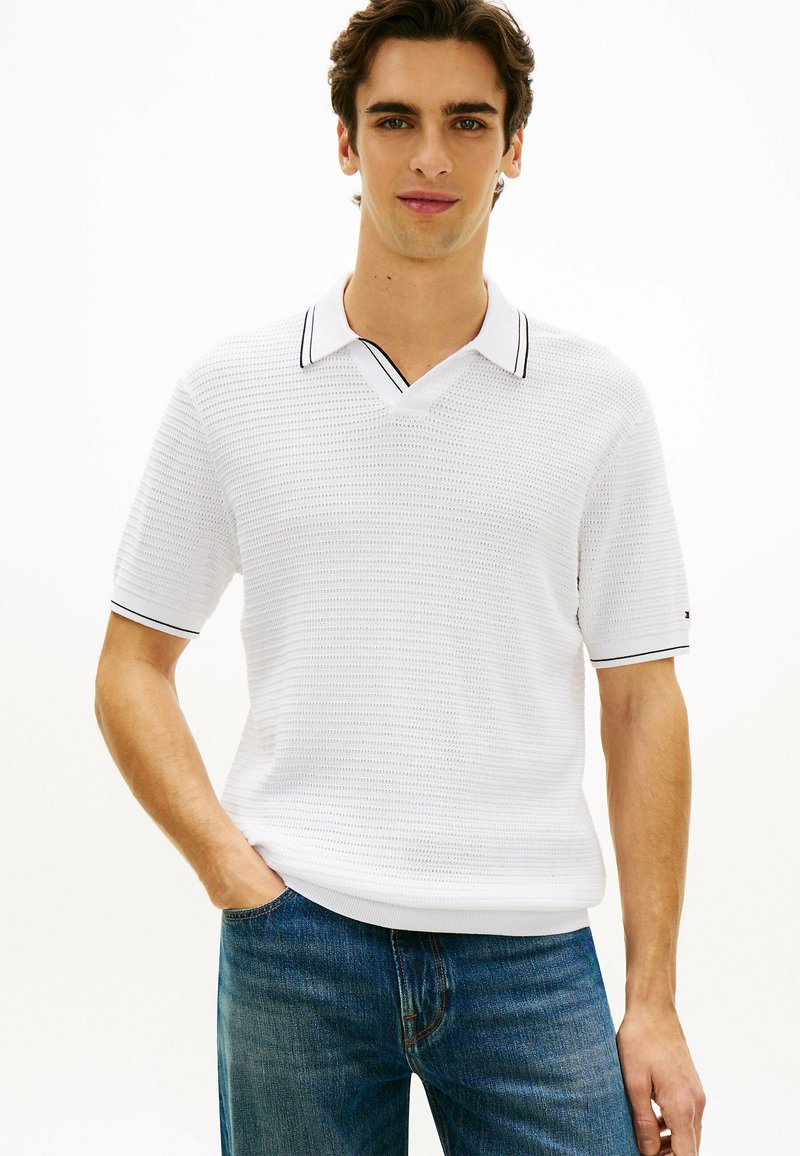 Jeune homme portant un polo en maille à manches courtes blanches avec des bordures noires, un jean bleu, debout avec une main dans la poche.