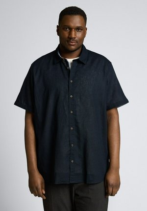 Mann trägt ein dunkelblaues kurzärmeliges Hemd mit Knöpfen und steht vor einem schlichten hellgrauen Hintergrund.