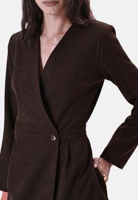 Donkerbruine wikkeljurk met een V-hals, lange mouwen, een knoopdetail op de taille en een gladde stoftextuur.
