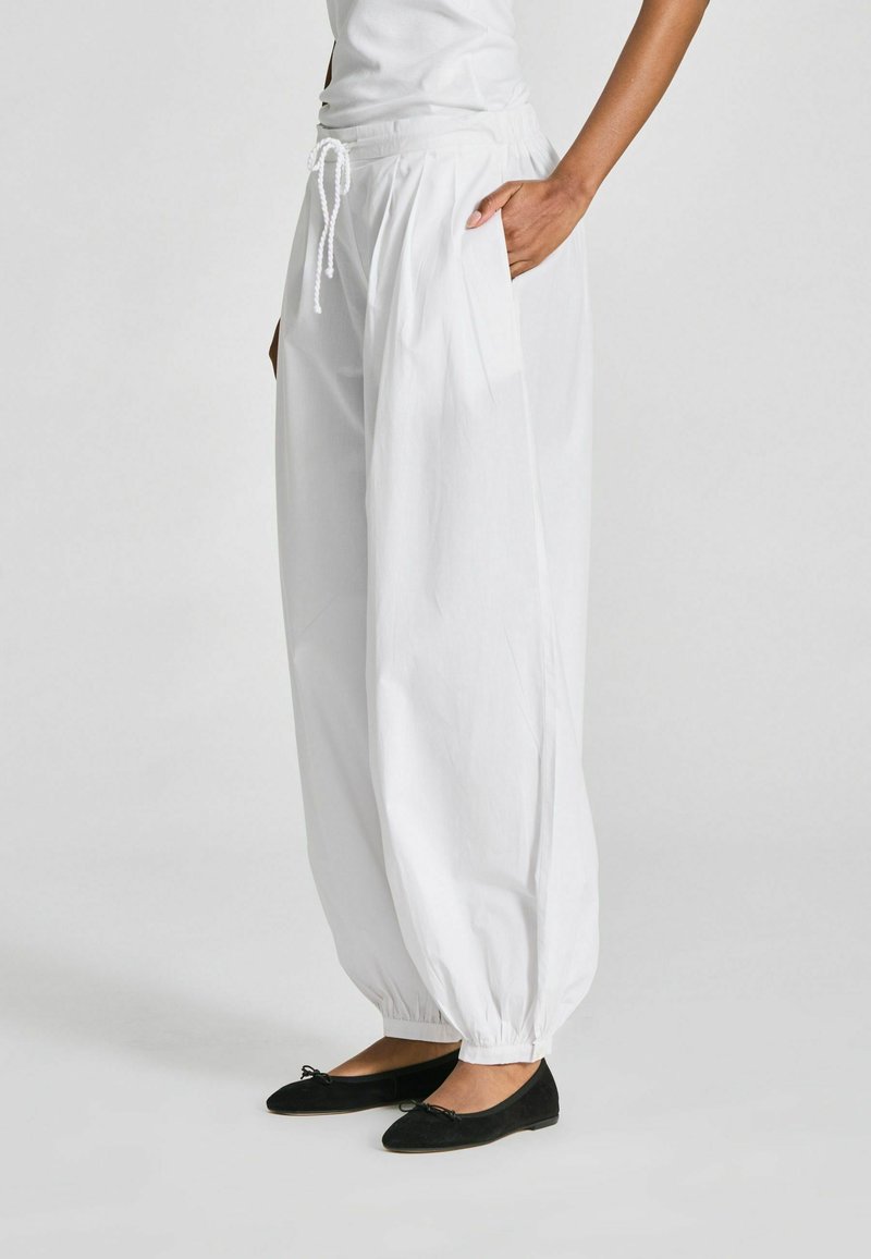 Personne portant un pantalon blanc ample à cordon avec chevilles élastiques et des ballerines noires, debout devant un fond uni.