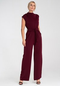 Bordowy kombinezon z krótkimi rękawami, paskiem w talii, szerokimi nogawkami i gładką teksturą. Posiada wysoki dekolt i minimalistyczną sylwetkę.
