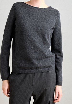 Personne portant un pull à manches longues gris foncé et un pantalon rayé sombre, sur un fond clair uni.