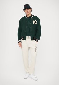 Πράσινο varsity μπουφάν από μάλλινη ύλη με κρεμ λωρίδες και λογότυπο NY, συνδυασμένο με κρεμ φόρμες που έχουν γκρι λογότυπο. Άσπρα sneakers ολοκληρώνουν το σύνολο.