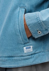 Χέρι στην τσέπη του ανάγλυφου ανοιχτό μπλε μπουφάν HUGO με μανίκι με μανσέτα και λεπτομέρεια με κουμπί, που δείχνει μέρος του γκρίζου παντελονιού από κάτω.