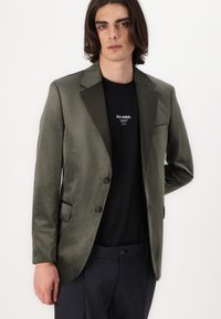 Olivgrüner Blazer mit satinierter Oberfläche, einreihigem Design, mit Reverskragen, zwei Knöpfen und einer Brusttasche, getragen über einem schwarzen Hemd.