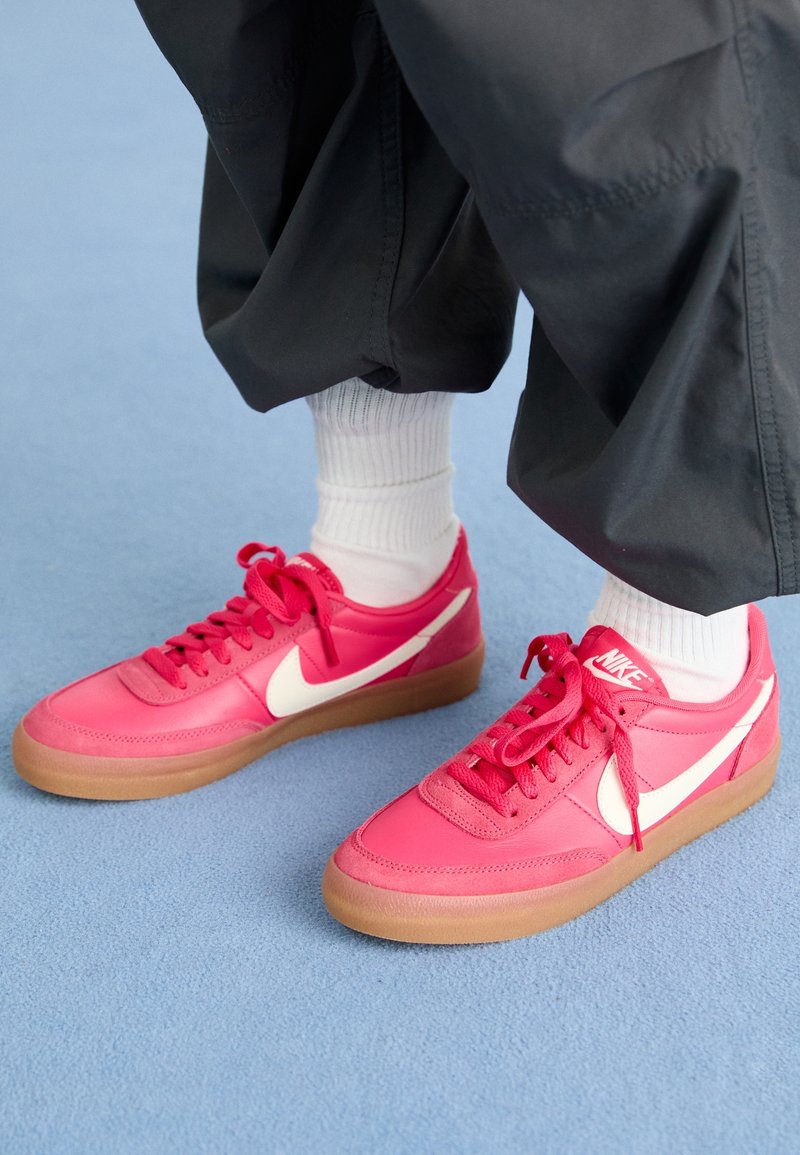 Pieds portant des chaussettes blanches et des baskets Nike roses vives à semelles en gomme, debout sur une surface bleu clair avec un pantalon noir.
