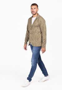 Blazer de color verde oliva, con textura, cuello con muesca, dos bolsillos frontales y cierre de un botón, combinado con jeans azules y zapatillas blancas.