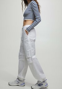 Vrouw in witte cargobroek, blauwe en witte gestreepte lange mouwen crop top, handen in de zakken, draagt witte en paarse sneakers, staat zijwaarts.