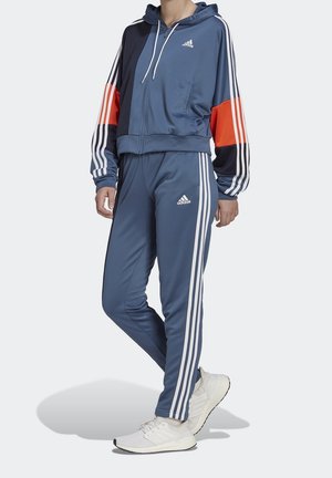 Person trägt einen blauen Adidas-Trainingsanzug mit weißen Streifen und orangen Akzenten, kombiniert mit weißen Adidas-Sneakern, stehend vor hellem Hintergrund.