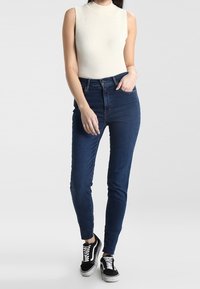 Gerippetes cremefarbenes ärmelloses Oberteil, kombiniert mit dunkelblauem Skinny-Jeans und schwarz-weißen Sneakers, die eine figurbetonte Silhouette und glatte Textur zeigen.