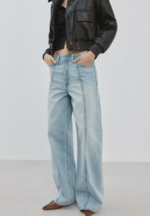 Persona vistiendo jeans anchos de pierna en azul claro con detalles de costura frontal, un top corto negro, chaqueta de cuero negra y zapatos puntiagudos marrones.