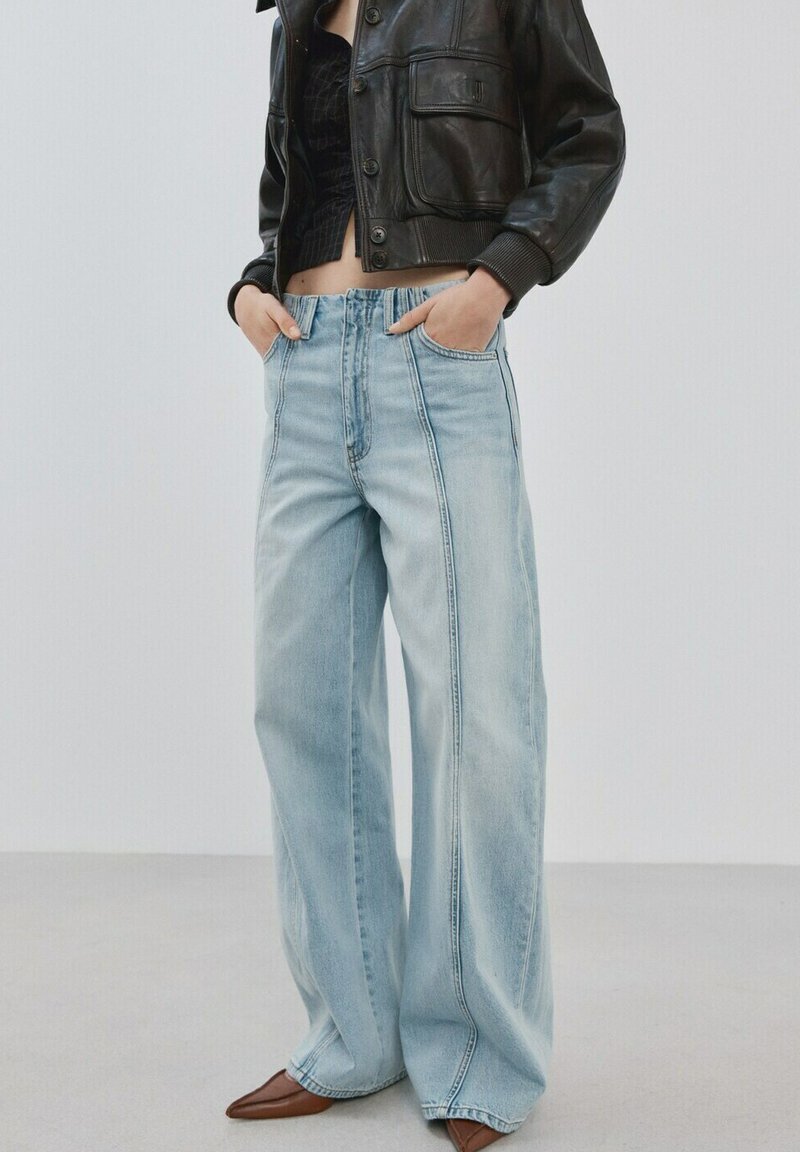 Person iført lyseblå jeans med vide ben og detalje med front-sømme, en kort sort top, sort læderjakke og brune spidse sko.