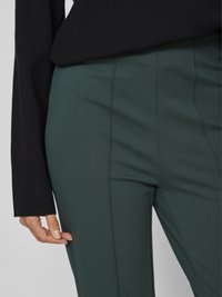 Pantalon tapered vert foncé avec une texture lisse et de subtils coutures verticales, associé à un pull noir à manches longues.