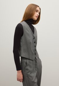 Gilet grigio con sei bottoni, motivo a zigzag, design cropped, abbinato a un dolcevita nero e pantaloni coordinati. Texture liscia.