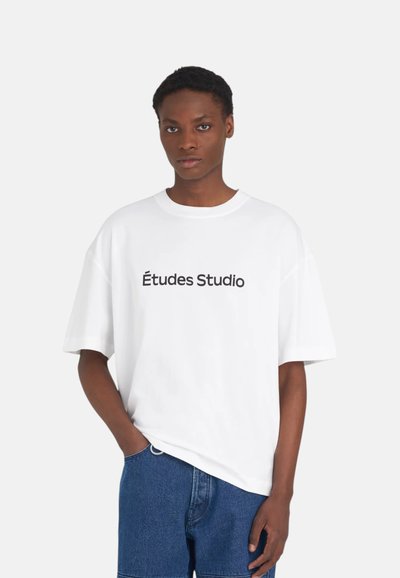 Personne portant un T-shirt blanc oversize avec l'inscription « Études Studio » en noir, associé à un short en denim bleu, debout devant un fond uni.