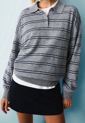 Femme portant un polo à manches longues gris à rayures par-dessus une chemise blanche, assorti d'une mini-jupe noire à fines rayures, devant un mur bleu clair.