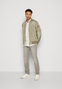 Light olive bomber jacket in smooth fabric, beige button-up shirt underneath, light gray jeans, and white sneakers with beige accents.