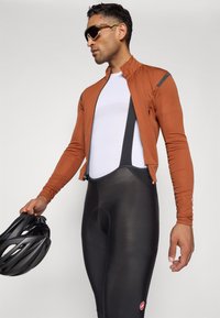 Sykkeldrakt som består av en brun langermet jakke, hvit tettsittende skjorte og svarte bib-shorts. Inkluderer en svart hjelm og solbriller.