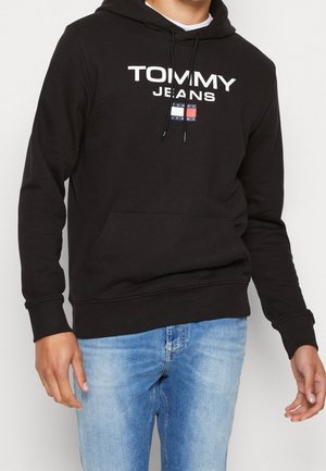 Persoon draagt een zwarte Tommy Jeans hoodie met wit logo en blauwe spijkerbroek, staand met ontspannen armen tegen een effen achtergrond.