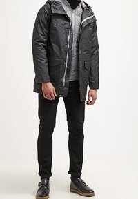 Homme portant des vestes noires et grises superposées, un pantalon noir et des chaussures noires, se tenant devant un fond blanc.