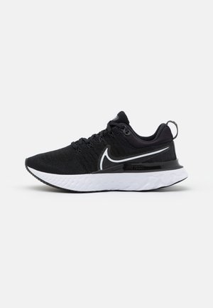 Sort og hvid atletisk løbesko med strikket overdel, sort Nike swoosh, hvid tekstureret sål og trækflig på hælen, sideudsigt på grå baggrund.