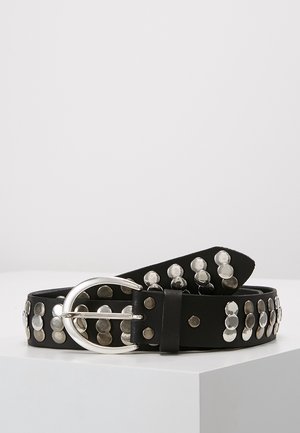 Zwarte leren riem versierd met meerdere rijen zilverkleurige ronde studs en een grote ovale zilveren gesp, opgerold geplaatst op een witte ondergrond.