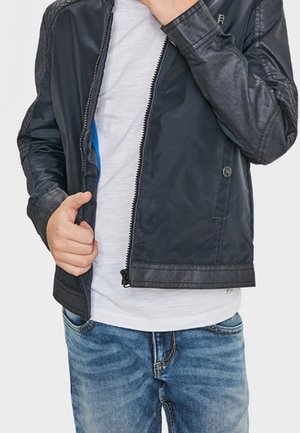 Persoon in een zwarte leren jas over een wit shirt en blauwe jeans, die met hun linke hand de rits van de jas aanpast.