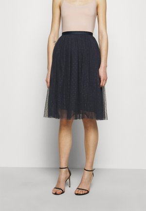 Czarna spódnica z tiulu z dopasowanym pasem, ozdobiona małymi groszkami, warstwowa dla objętości, do kolan, w połączeniu z czarnymi sandałkami na paskach.