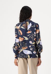 Blusa floreale navy con maniche a sbuffo, scollatura arricciata e schiena con apertura a chiave. Presenta vivaci motivi floreali nei colori arancione, viola e crema.