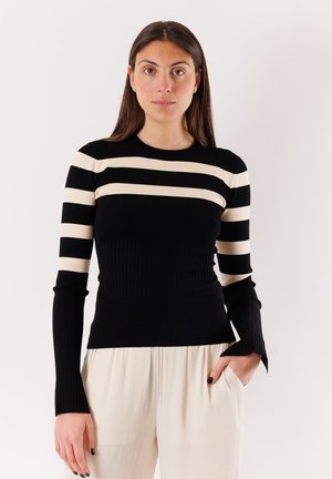 Donna con lunghi capelli scuri che indossa un maglione nero a strisce bianche e pantaloni chiari, in piedi davanti a uno sfondo semplice.