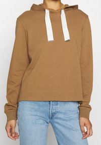 Felpa con cappuccio marrone con coulisse bianche, caratterizzata da una vestibilità comoda e polsini a coste. Indossata sopra jeans azzurri.