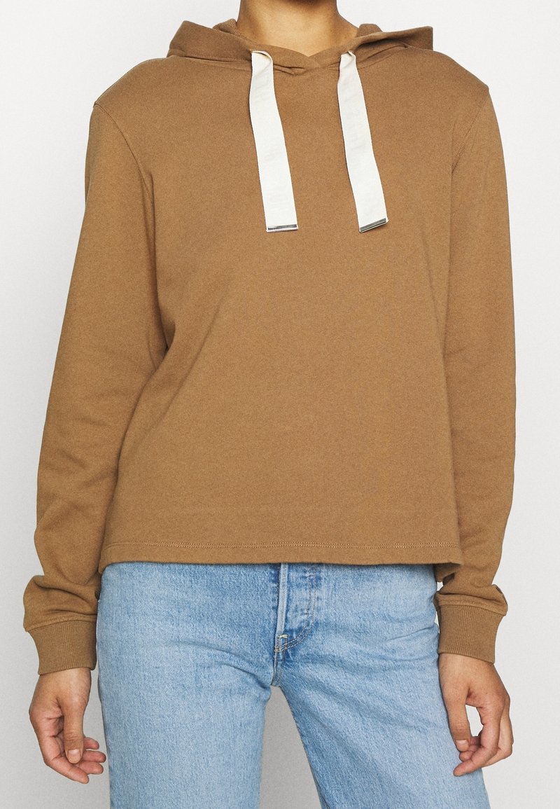 Felpa con cappuccio marrone con coulisse bianche, caratterizzata da una vestibilità comoda e polsini a coste. Indossata sopra jeans azzurri.