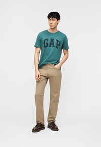 Νέος άνδρας standing με το ένα χέρι στην τσέπη, φοράει μπλε-πρασινό μπλουζάκι GAP, μπεζ παντελόνι και καφέ παπούτσια σε απλό λευκό φόντο.