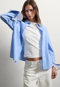 Mujer con cabello castaño que lleva una blusa blanca y pantalones, posando con un brazo doblado detrás de su cabeza y una camisa a rayas azul desabotonada.