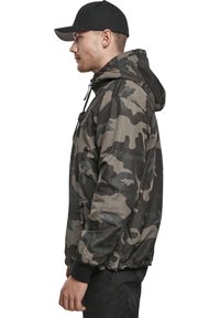 Chaqueta de camuflaje en tonos de verde y gris. Cuenta con capucha, puños elásticos y un bolsillo con cremallera. El material es suave y ligero.