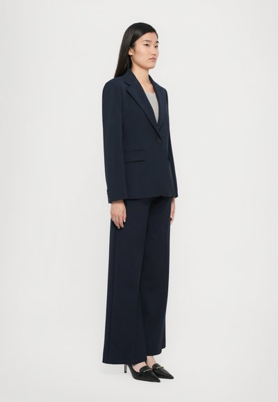 Abito blu navy su misura con blazer strutturato e pantaloni a gamba larga, realizzato in tessuto morbido. Tacchi a punta neri aggiungono contrasto.