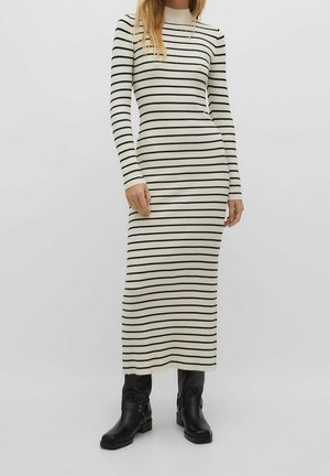 Woman wearing a long-sleeve, white ribbed knit dress with black horizontal stripes and black mid-calf boots, standing against a plain background.