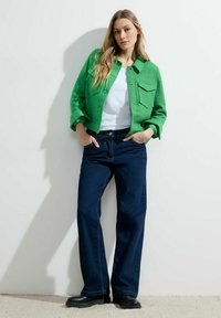 Chaqueta de mezcla de lana verde con dos bolsillos frontales, llevada sobre una camisa blanca. Combinada con jeans de pierna ancha azul oscuro y botas negras de tobillo.