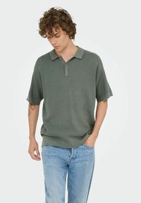 Strikket grøn polo skjorte med en struktureret overflade, korte raglanærmer og en placket med to knapper. Sammen med lyseblå denim jeans.