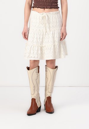 Femme portant une jupe blanche ajourée mi-longue à œillets avec une taille à cordon et des bottes de cowboy beige et marron.