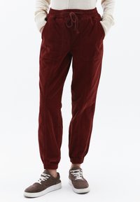 Červené kordurové jogger kalhoty s elastickým pasem a šňůrkou, se bočními kapsami a žebrovanými manžetami, v kombinaci s hnědými teniskami.