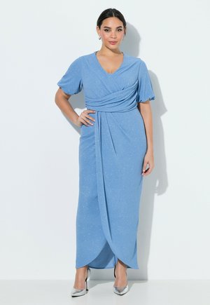 Femme debout en robe portefeuille bleu clair scintillante à manches courtes et talons hauts argentés sur un fond blanc uni.