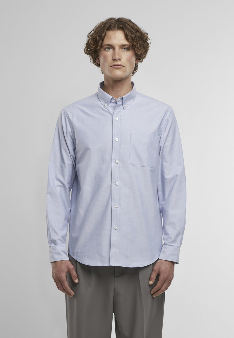 Jeune homme aux cheveux bouclés, portant une chemise boutonnée bleu clair et un pantalon gris, debout devant un fond uni gris clair.