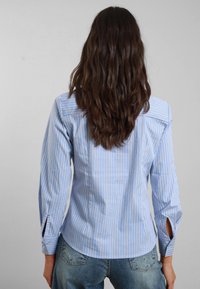 Camicetta a righe in azzurro chiaro e bianco, con maniche lunghe e fessure laterali, vestibilità sartoriale e orlo leggermente curvato.