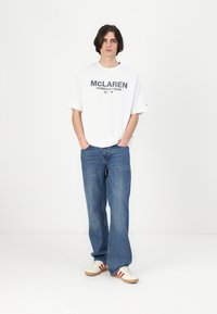 Balta grafiskā T-kreklu ar tekstu "McLaren Formula 1 Team", apvienots ar ziliem platām kājām džinsiem un brūniem sporta apaviem ar sarkaniem svītrām.