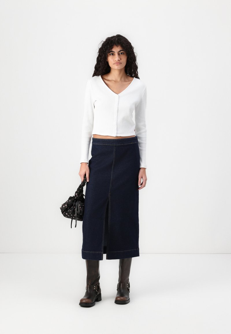 Weißes, geripptes Langarm-Crop-Top mit V-Ausschnitt; dunkelblaue Jeans-Maxirock mit Schlitz vorne; kombiniert mit braunen Stiefeletten und einer schwarzen Tasche.